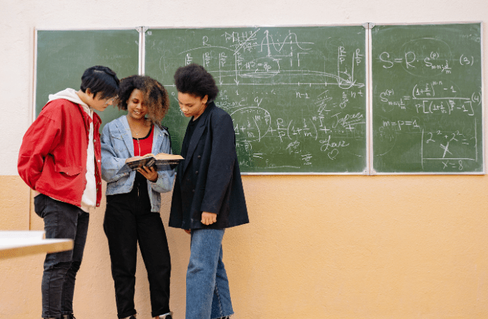 Tre studenti che guardano un libro davanti a una lavagna piena di numeri, scena scolastica legata alla plusdotazione scuole superiori